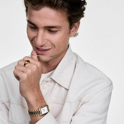 Man wearing a gold Daniel Wellington Tide mesh necklace and matching gold watch, styled with a casual white jacket.